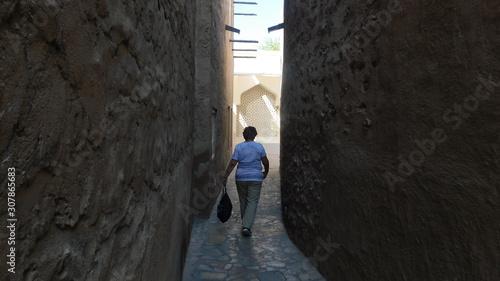 Narrow dark  street lined  with high  stone  walls in Arabic style with a  woman strolling on a sunny summer day
