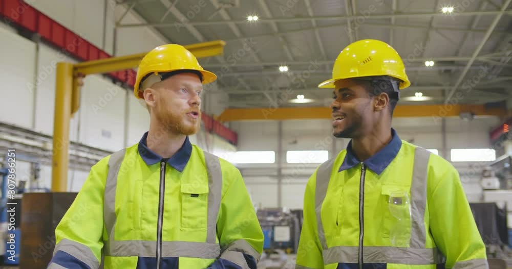 Two factory workers walking with discussion in industrial heavy industry enterprise background. Foremen in helmets and uniform walking through factory facility Two factory workers walking with discussion in industrial heavy industry enterprise background. Foremen in helmets and uniform walking through factory facility