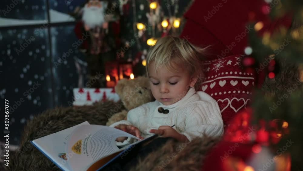 Cute toddler boy, sitting around christmas tree in cozy chair, reading book Cute toddler boy, sitting around christmas tree in cozy chair, reading book