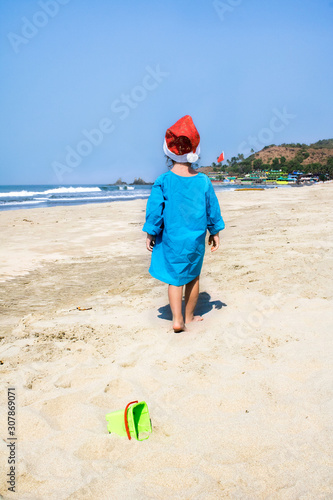 Little child in a Santa hat walks on a tropical beach.