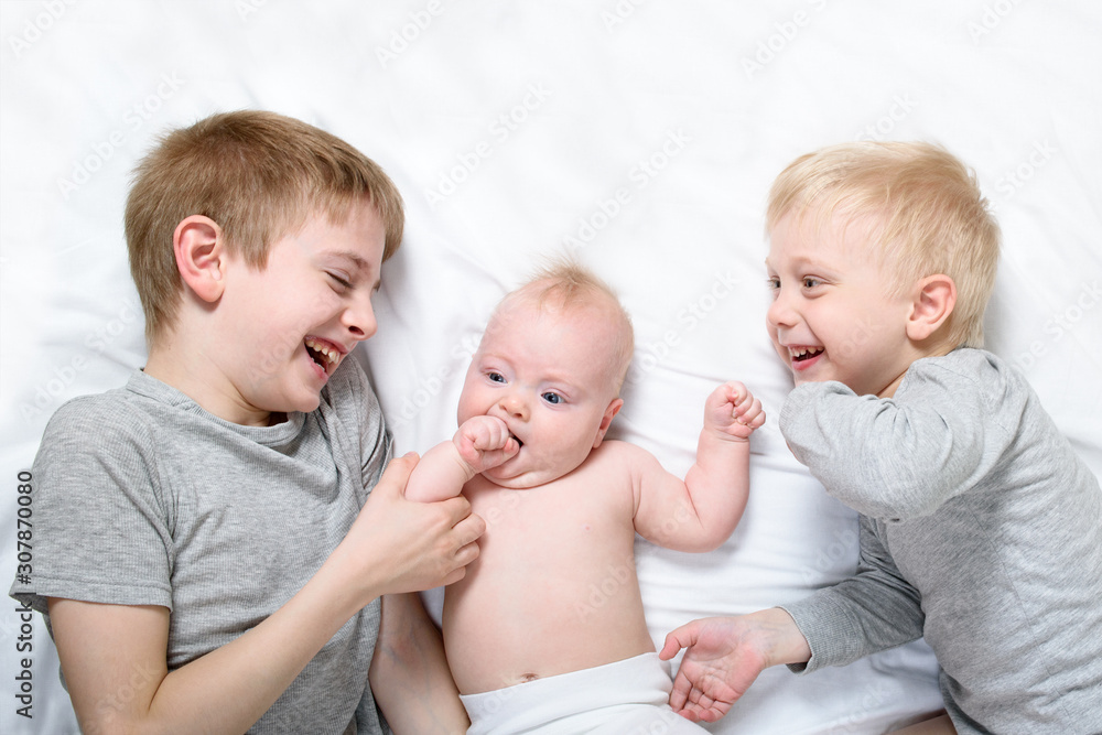 Two older brothers play and laugh with the youngest baby in a white bed. Happy childhood, big family Two older brothers play and laugh with the youngest baby in a white bed. Happy childhood, big family