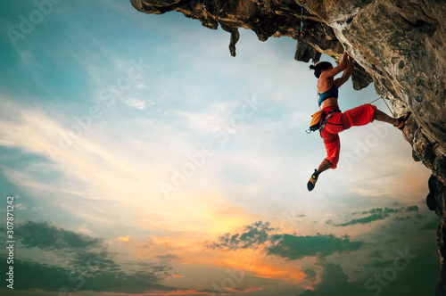 Fototapeta Athletic Woman climbing on overhanging cliff rock with sunset sky background