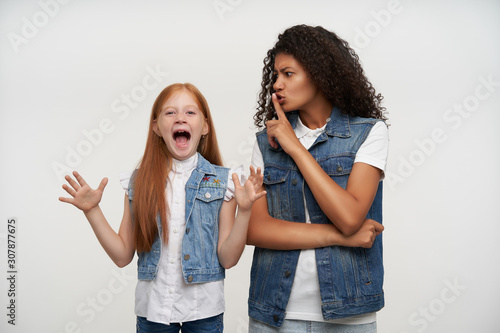 Studio photo of young curly brunette lady with dark skin raising hand in hush gesture and trying to calm down energized lovely redhead little girl  isolated over white background