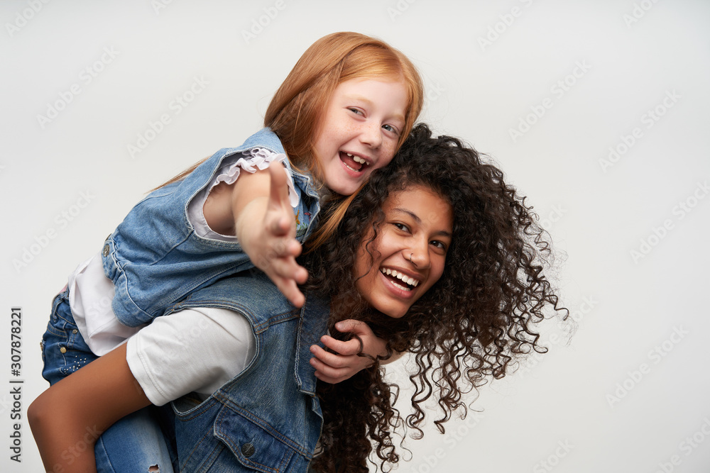 Side view of joyful pretty young dark skinned curly brunette woman riding on her back cheerful cute redhead female kid, looking happily to camera and smiling broadly, isolated over white background
