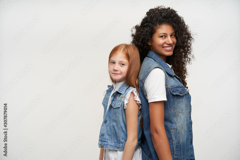 Couple of young positive attractive ladies looking cheerfully to camera with charming smiles, standing back to each other while posing over white background with hands along bodies Couple of young positive attractive ladies looking cheerfully to camera with charming smiles, standing back to each other while posing over white background with hands along bodies