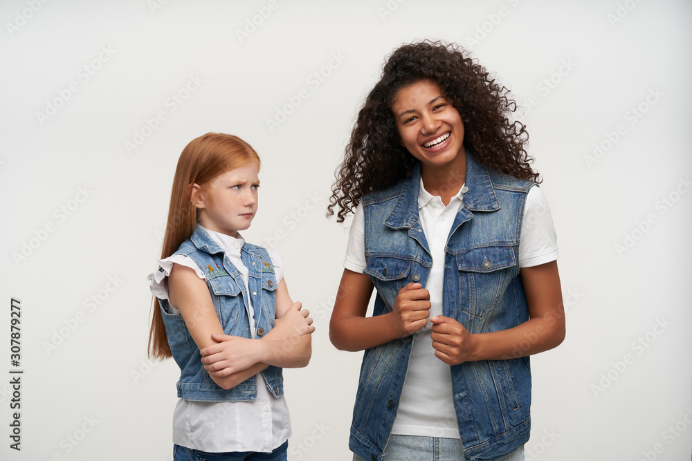 Displeased foxy long haired female kid folding her hands and frowning eyebrows while looking discontentedly at her young dark skinned happy female friend with brown curly hair Displeased foxy long haired female kid folding her hands and frowning eyebrows while looking discontentedly at her young dark skinned happy female friend with brown curly hair