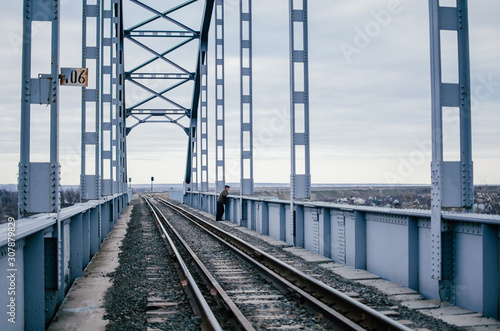 Man stands bored on rails of an old railway bridge, sadly waiting for a meeting. Industrial landscape. Big iron bridge on a cloudy day. The concept of separation, meeting, depression. Copy space