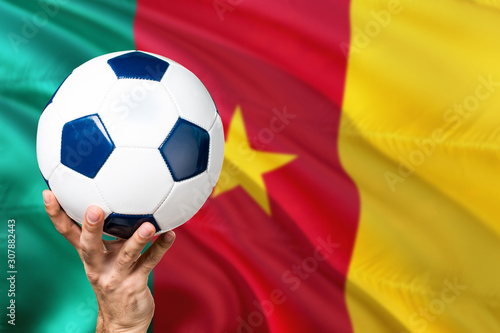 Cameroon soccer concept. National team player hand holding soccer ball with country flag background. Copy space for text.