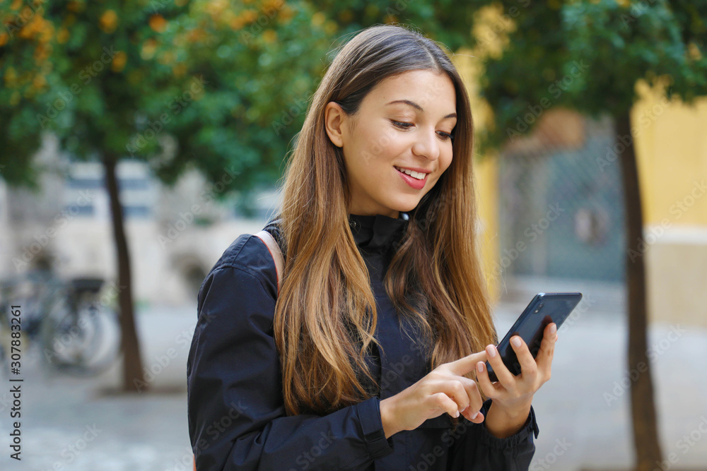 Portrait of a beautiful smiling woman using a mobile phone in city street with tangerine trees on the background.