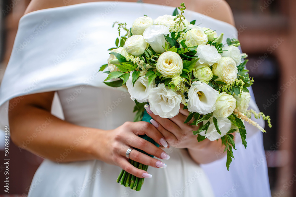 White wedding bouquet of different flowers and dried flowers in the hands of the bride on a green background. Hands of the bride and dress are artistically blurred against the backdrop White wedding bouquet of different flowers and dried flowers in the hands of the bride on a green background. Hands of the bride and dress are artistically blurred against the backdrop