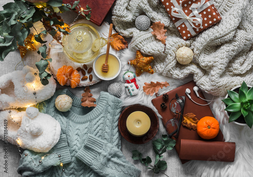 Christmas mood cozy home still life. Knitted plaid, sweater, books, green tea with honey, christmas toys, light garland, candle, soft slippers on the fluffy carpet, top view. Flat lay