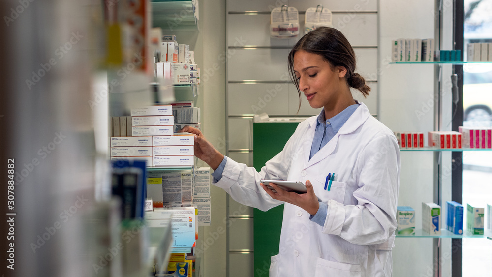 An young female pharmacist consultant is checking with a tablet medicines on the shelf of drug store. 