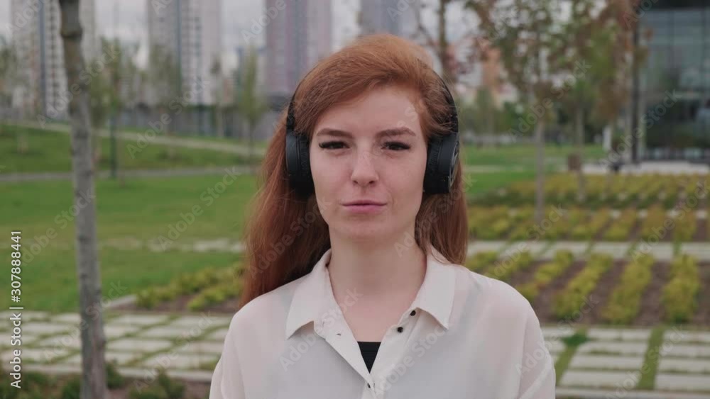 Smiling young woman with red hair standing and listening to music with big wireless headphones in summer city street. Handheld real time medium shot Smiling young woman with red hair standing and listening to music with big wireless headphones in summer city street. Handheld real time medium shot