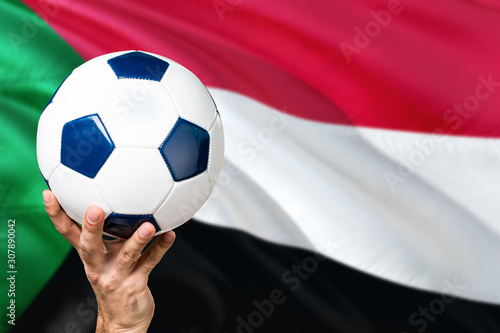 Sudan soccer concept. National team player hand holding soccer ball with country flag background. Copy space for text.