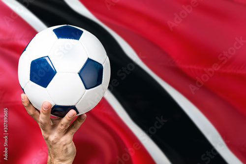 Trinidad And Tobago soccer concept. National team player hand holding soccer ball with country flag background. Copy space for text.