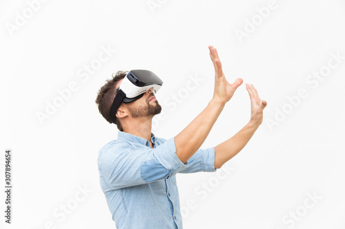 Side of bearded guy wearing VR goggles, touching air. Young man in virtual reality headset standing isolated over white background. VR game concept
