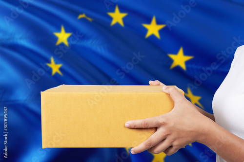 European Union delivery service. International shipment theme. Woman courier hand holding brown box isolated on national flag background.