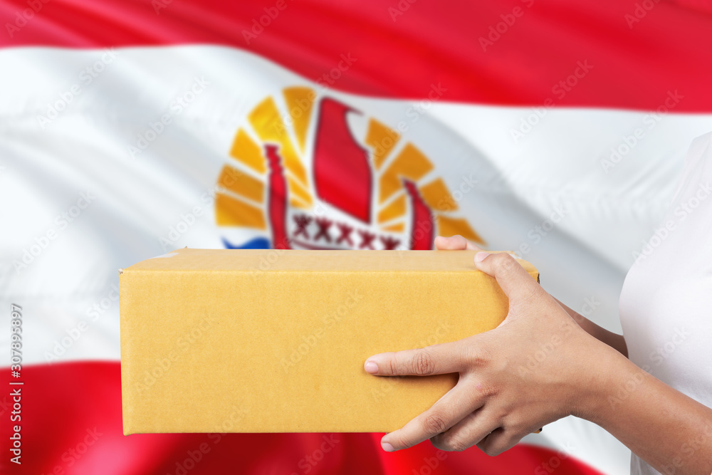 Fototapeta premium French Polynesia delivery service. International shipment theme. Woman courier hand holding brown box isolated on national flag background.