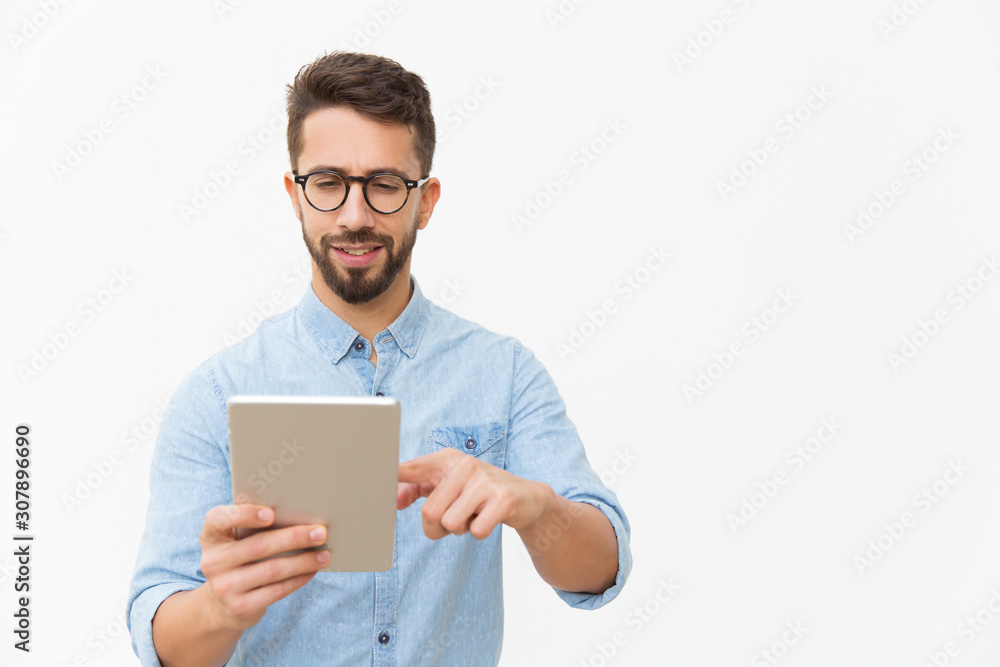Focused positive guy watching content on tablet. Handsome young man in casual shirt and glasses standing isolated over white background. Digital technology concept Focused positive guy watching content on tablet. Handsome young man in casual shirt and glasses standing isolated over white background. Digital technology concept