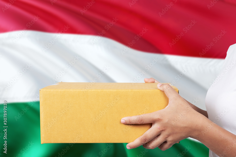 Obraz premium Hungary delivery service. International shipment theme. Woman courier hand holding brown box isolated on national flag background.