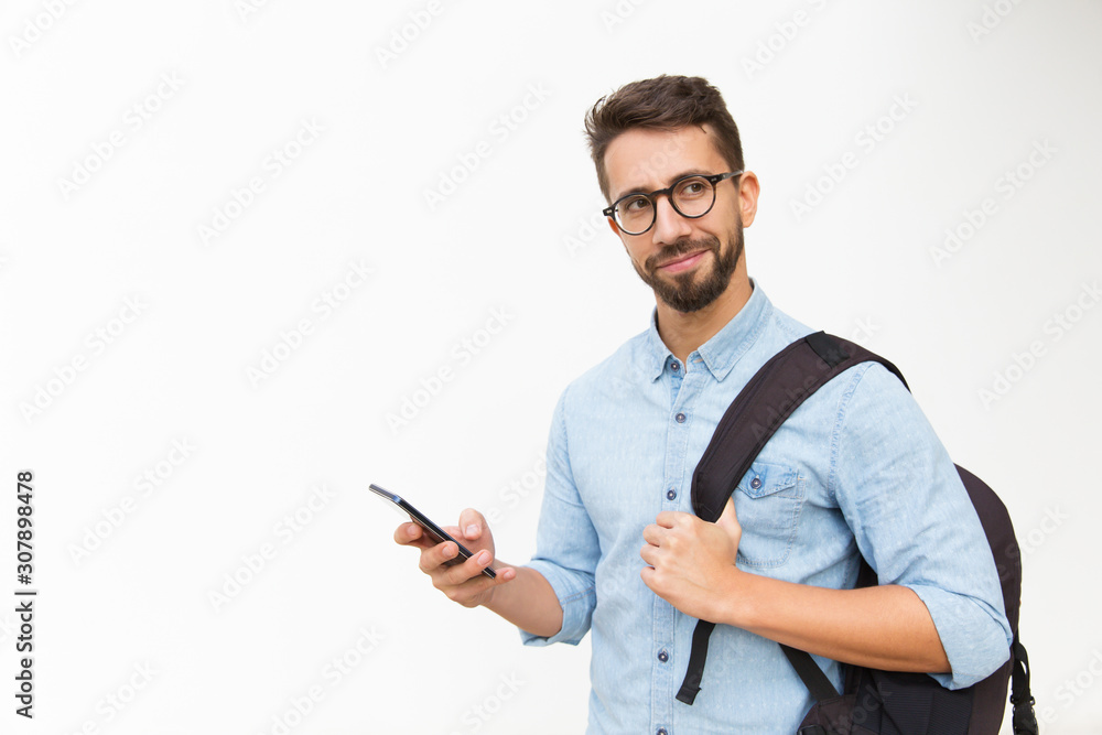 © Mangostar - Positive male tourist with backpack using mobile phone, looking away. Handsome young man in casual shirt and glasses standing isolated over white background. Communication or travel concept