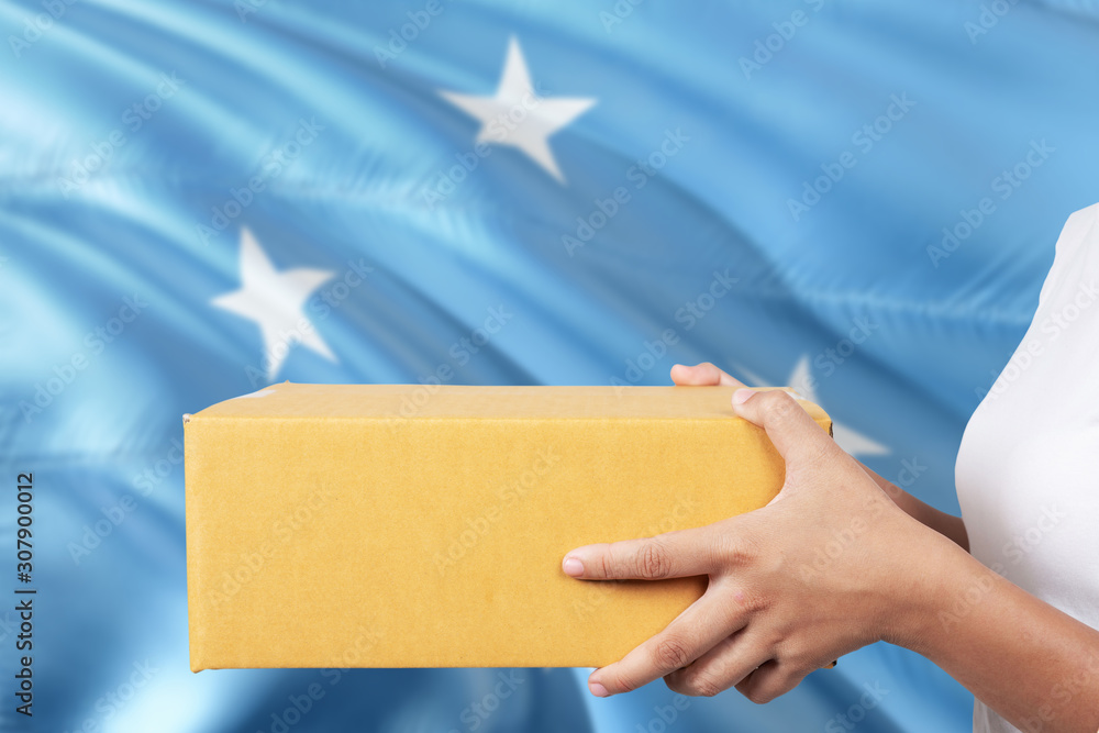 Obraz premium Micronesia delivery service. International shipment theme. Woman courier hand holding brown box isolated on national flag background.