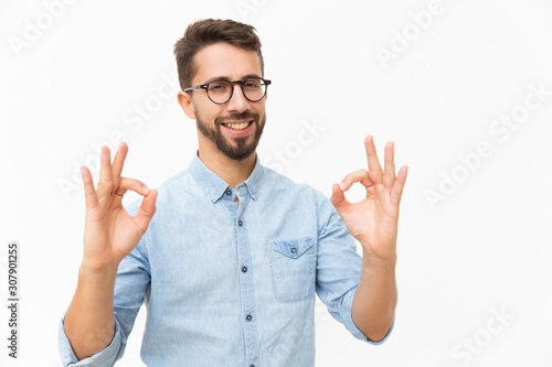 Happy joyful guy showing OK gesture with both hands. Handsome young man in casual shirt and glasses standing isolated over white background. Customer satisfaction concept photo