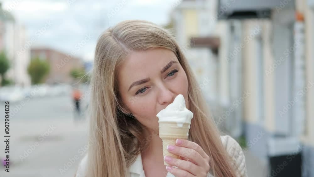 A young attractive blonde woman eats a white ice cream horn in a waffle cup on the street. Summer day in the city. A young attractive blonde woman eats a white ice cream horn in a waffle cup on the street. Summer day in the city.