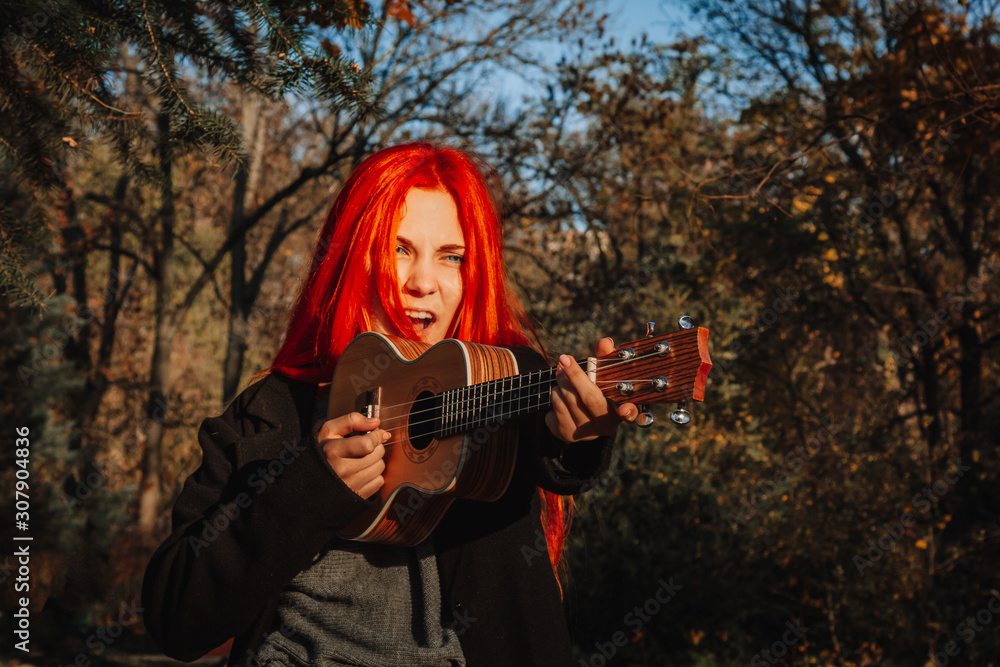 Red-haired girl with long hair plays on the ukulele in the park. School, music education concept, student learns to play the string instrument. Hands of a musician, classical, melody, creativity.