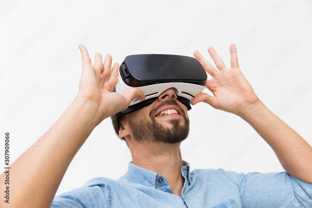 Smiling male user wearing VR glasses, adjusting device, turning face up. Young man in virtual reality headset standing isolated over white background. VR experience concept Smiling male user wearing VR glasses, adjusting device, turning face up. Young man in virtual reality headset standing isolated over white background. VR experience concept