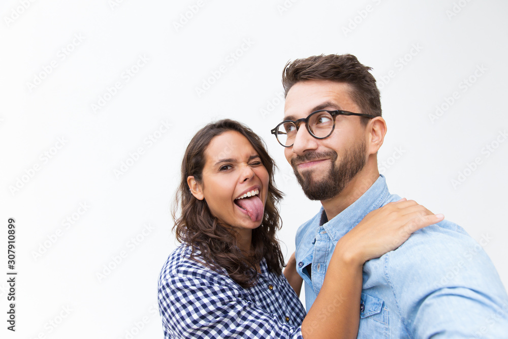 Joyful funny couple grimacing for selfie, smiling and sticking tongue out. Young woman in casual and man in glasses standing isolated over white background. Couple having fun concept Joyful funny couple grimacing for selfie, smiling and sticking tongue out. Young woman in casual and man in glasses standing isolated over white background. Couple having fun concept