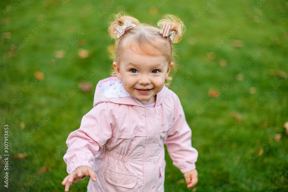 Little girl in cute pink jacket standing in the autumn park in the warm weather