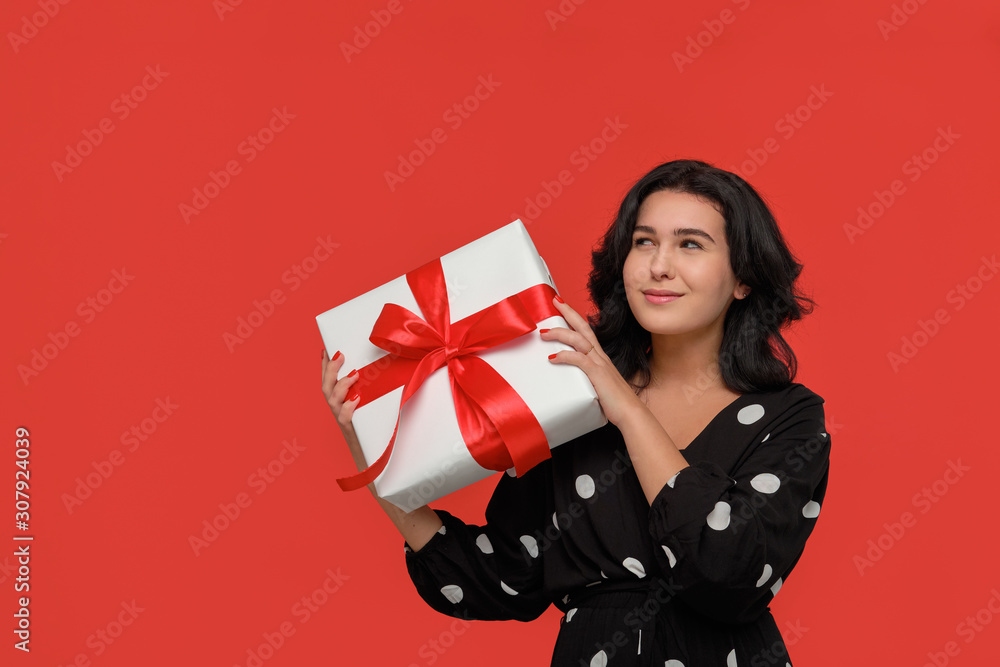 Brunette woman in a black dress smiling holding Christmas giftbox of white color with red ribbon. Brunette woman in a black dress smiling holding Christmas giftbox of white color with red ribbon.