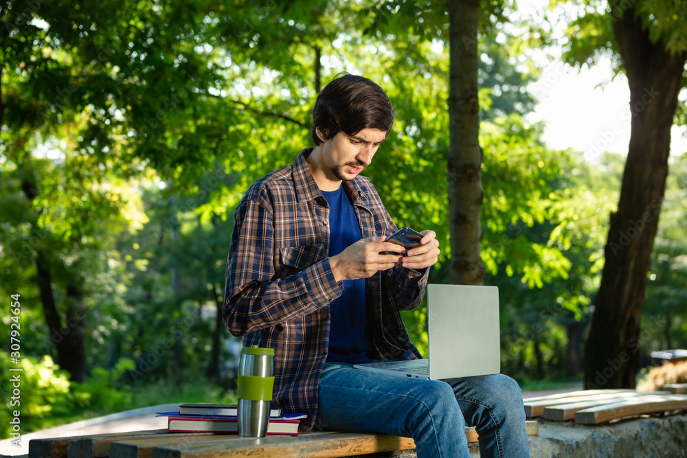 Young brunet freelancer with beard and mustache sitting with a laptop and coffee in a park. Young brunet freelancer with beard and mustache sitting with a laptop and coffee in a park.