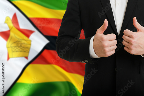 Zimbabwe businessman showing thumbs up behind country flag with copy space. Successful international relations and agreement concept.