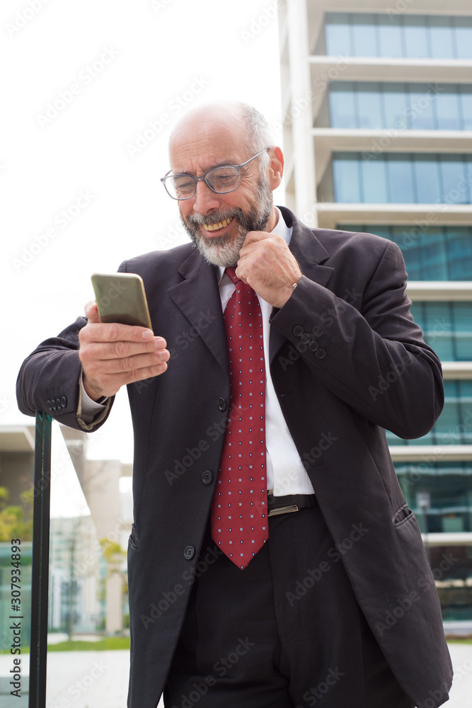 Cheerful businessman using smartphone on street. Low angle view of happy mature bearded businessman in eyeglasses standing outdoors and using mobile phone. Business and technology concept Cheerful businessman using smartphone on street. Low angle view of happy mature bearded businessman in eyeglasses standing outdoors and using mobile phone. Business and technology concept