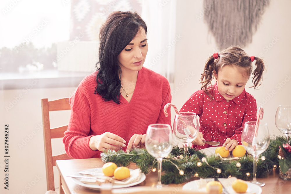 Cheerful mother and her daughter laughing, having fun with Christmas tree and star shaped cookies at the kitchen table. They are in red clothes wearing reindeer antlers and Santa hat headbands. Cheerful mother and her daughter laughing, having fun with Christmas tree and star shaped cookies at the kitchen table. They are in red clothes wearing reindeer antlers and Santa hat headbands.