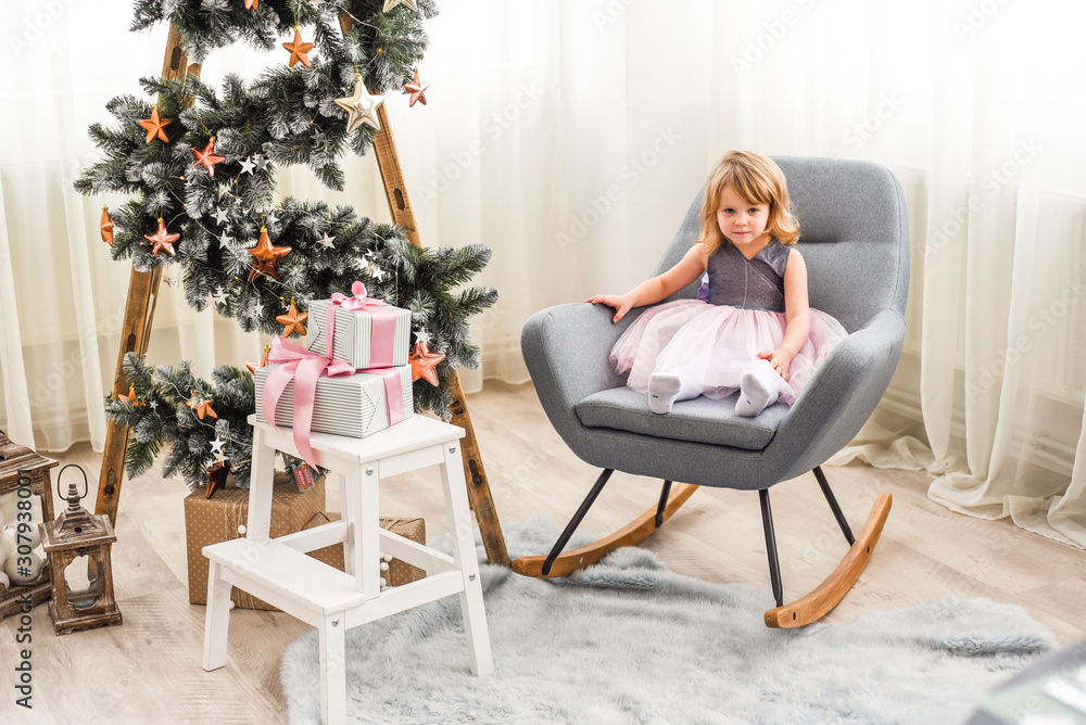 beautiful girl in a pink dress sitting on a chair near a Christmas tree beautiful girl in a pink dress sitting on a chair near a Christmas tree