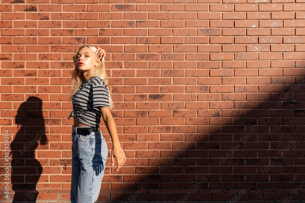 Attractive blonde teen girl in grey striped y-shirt and jeans touching her hair and lookinf at the camera isolated over red brick wall