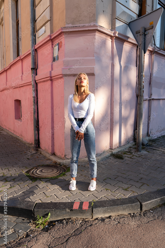 Street style portrait of the beautiful blonde teen girl in white top and blue jeans stands near the corner of the pink old house