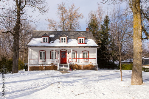 Canvas Print Beautiful white french-style ancestral clapboard house with red trimmed windows and door and grey shingled roof seen during winter, Ste