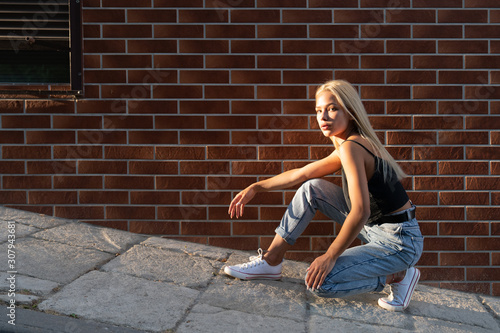 Trendy teen blonde girl in casual clothes crouched near the red brick wall, sun rays are falling on her face