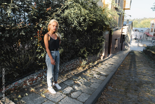 Attarctive calmy blonde girl in casual clothes stands near the green fence among the old city street photo