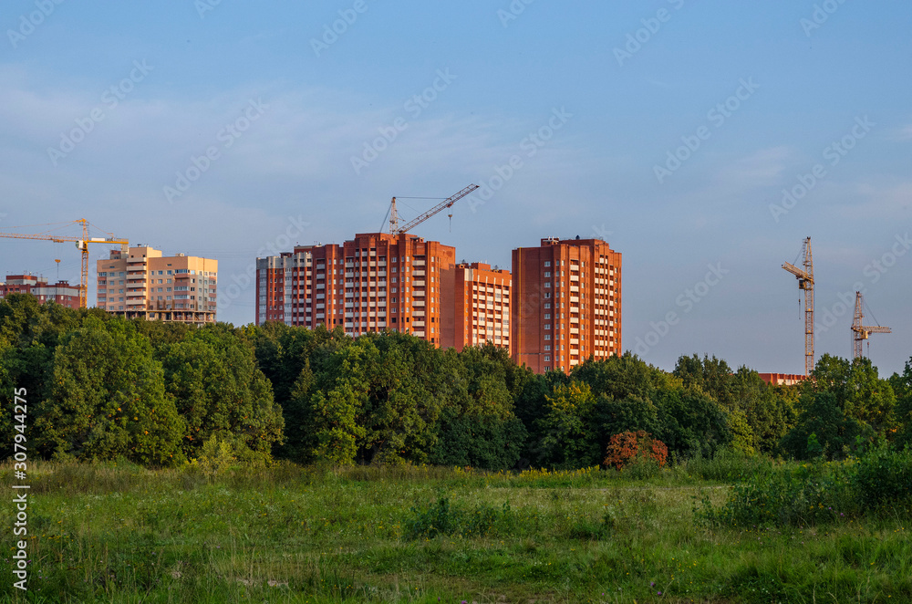 Tall tower crane near a multi-storey modern house under construction near a green forest against the background of finished houses.
