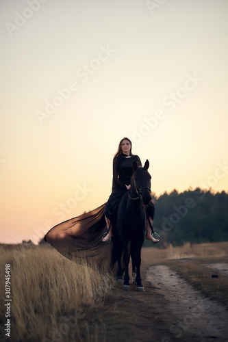 cheerful smiling young beautiful girl in a light flying black dress riding a black horse rides on the field against the background of the forest and sunset © MikeFrame