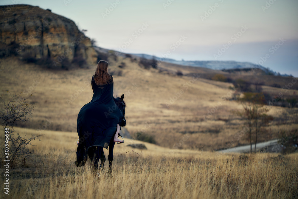 Cheerful smiling young beautiful girl in a light flying black dress riding a black horse stands with his back and looks at the horizon