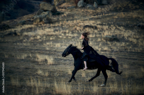 cheerful young beautiful girl in a light flying black dress riding on a black horse gallops on a field against the backdrop of mountains and sunset © MikeFrame