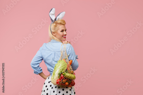 Happy Easter. Young pin up woman with bunny ears hold a string bag full of organic vegetables on pink background. Happy face. Emty space for text