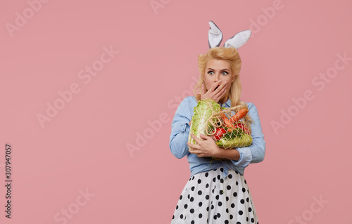 Upset pin up woman with easter bunny ears hold a string bag full of organic vegetables on pink background.
