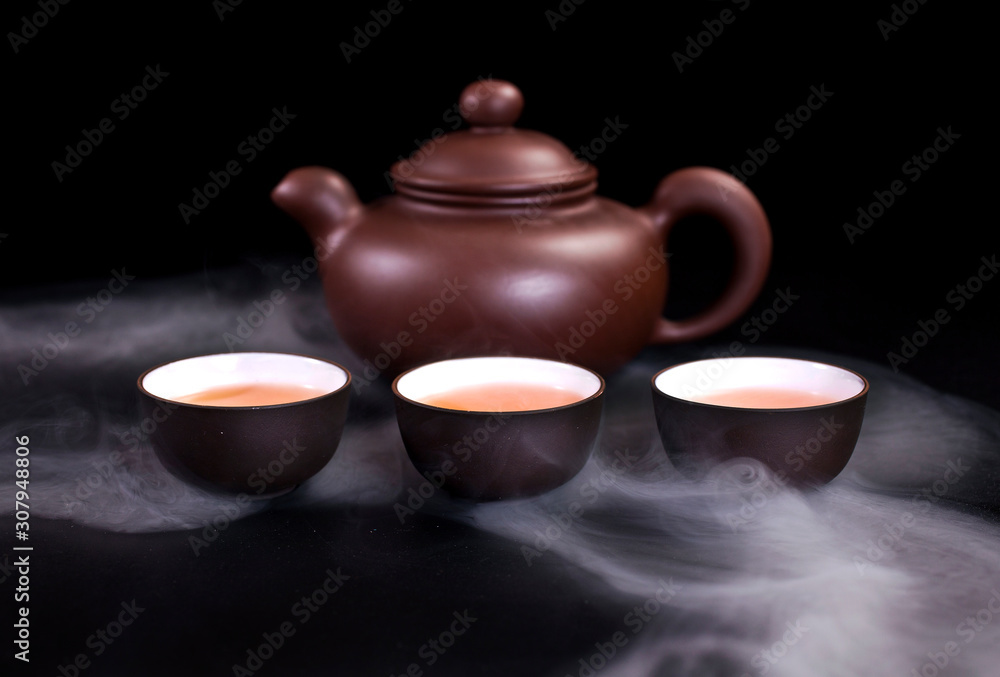 Asian tea ceremony. Chinese clay teapot and ceramic tea cups with vapor on a black background. Asian tea ceremony. Chinese clay teapot and ceramic tea cups with vapor on a black background.